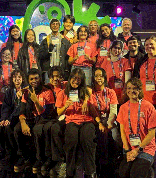 A diverse group of people pose together, most wearing matching red shirts, smiling and holding badges.