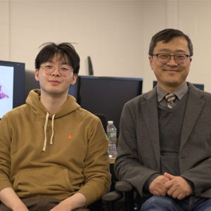Student and professor sitting together in front of computers smiling at the camera.