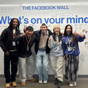 A group of five smiling people stand in front of "The Facebook Wall" with "What's on your mind" text. They pose happily, some giving peace signs.