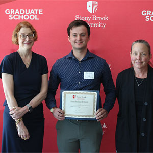 Three people pose in front of a red Stony Brook University Graduate School backdrop, with the person in the center holding a framed certificate.