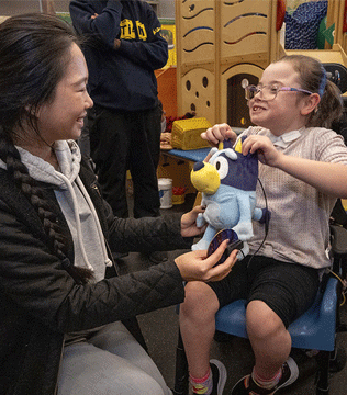 A young girl in a wheelchair joyfully adjusts a plush toy with student.