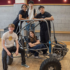 A diverse group of five students smiles while posing with a rugged off-road vehicle frame.