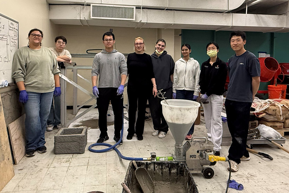 A group of eight individuals posing in an engineering workshop next to a concrete mixing pump and a 3D-printed concrete structure.