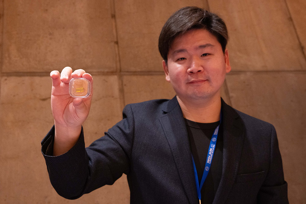 Man in a black blazer holds a clear square container with a golden object inside, standing in front of a textured concrete wall.