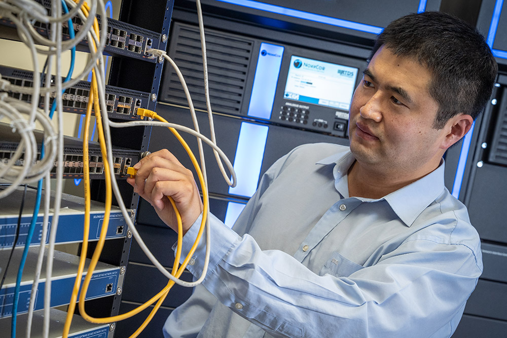 A man in a light blue shirt connects a yellow ethernet cable to a network switch in a server rack, surrounded by various tangled cables and illuminated by blue lighting.