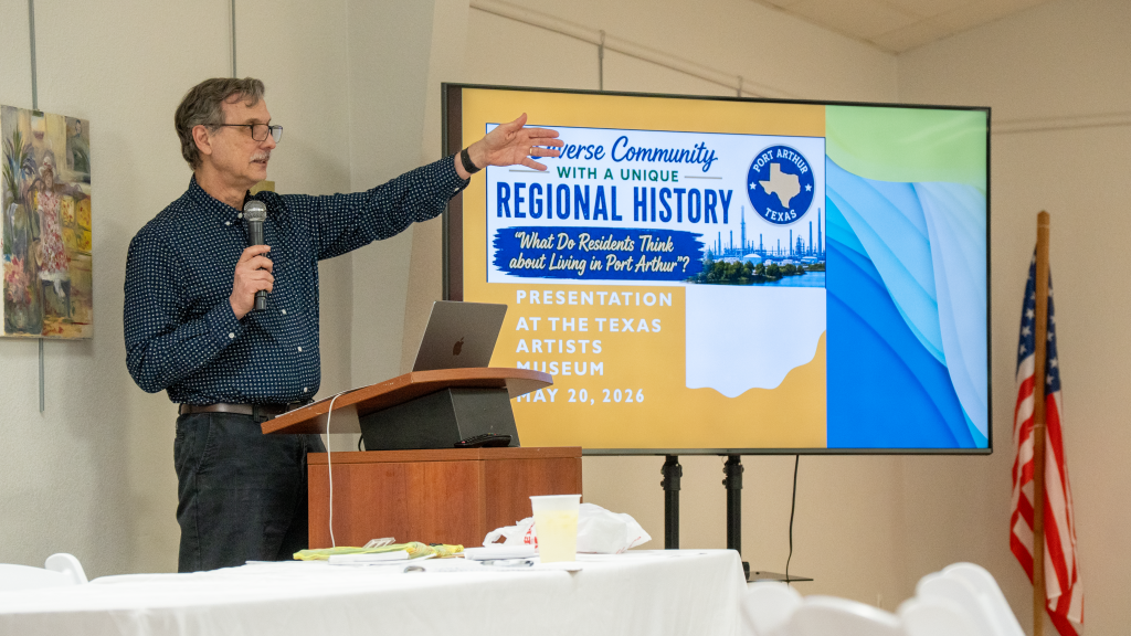 Dr. Christopher Sellers stands behind a podium presenting research findings displayed on a TV screen to his left