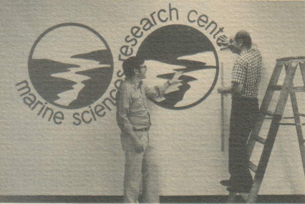 MSRC’s logo is prominently displayed in Endeavour Hall. In this picture from 1976, former MSRC director J. R. Schubel, left, enlisted the services of his father-in-law, Dr. Marion Hostetler, Montpelier, Ohio, in the execution of the design. The logo consists of reduplicated circles depicting an estuary emptying into the ocean. The circle on the left is green, representing land and vegetation, while the one on the right is blue, representing the estuary and the sea. The logo graphically depicts MSRC’s commitment to the coastal environment.
