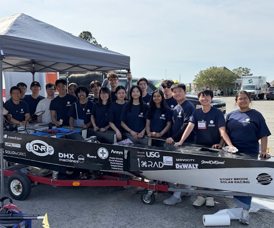 A group of students from Stony Brook Solar Racing stand behind their solar-powered vehicle, which is displayed on a trailer under a grey canopy.