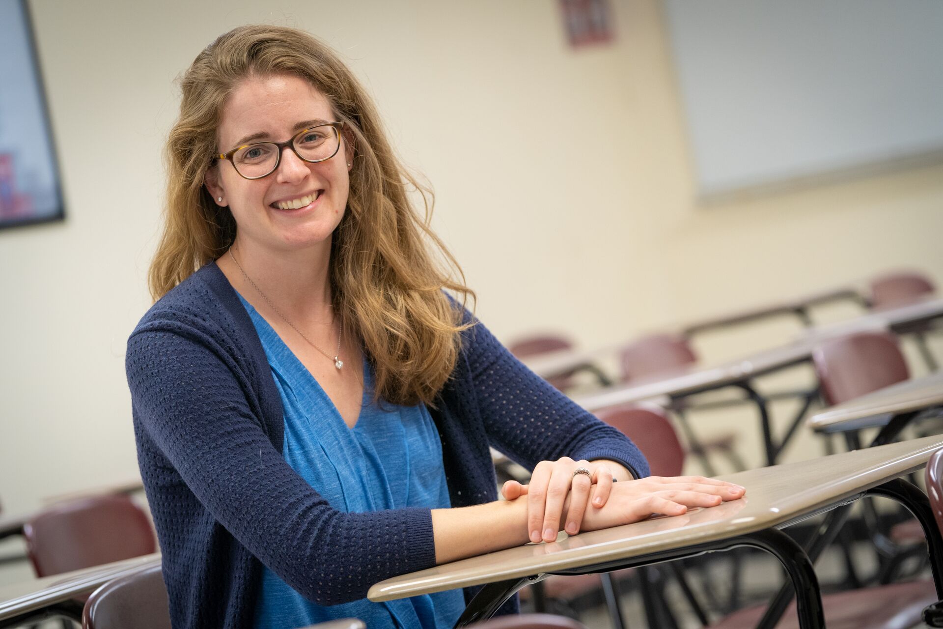 Caucasian woman with long blonde hair wearing dark blue cardigan over lighter blue shirt, sitting in classroom desk