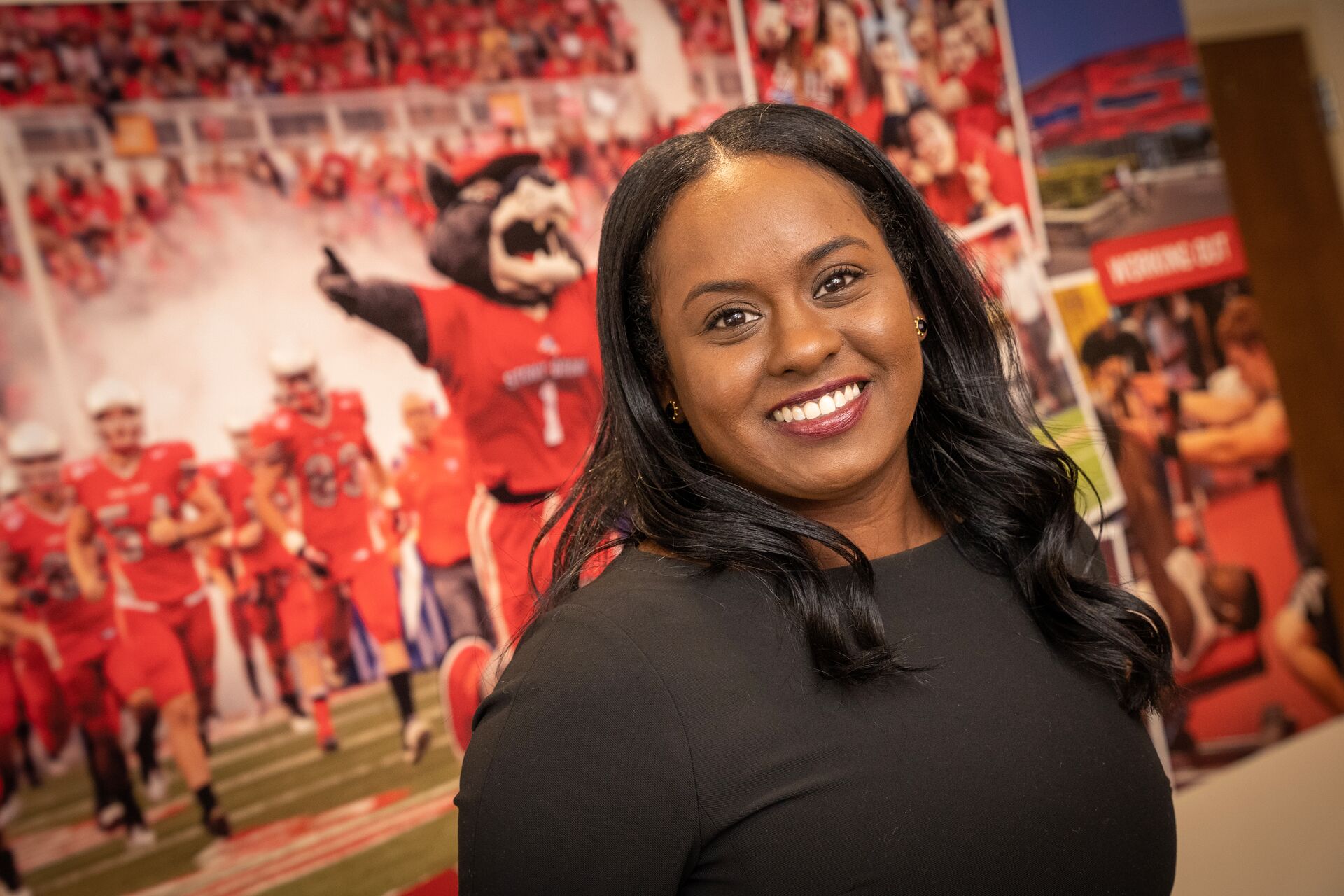 African American woman with long curled hair and black shirt, standing in front of wall with graphic of Wolfie and the football team