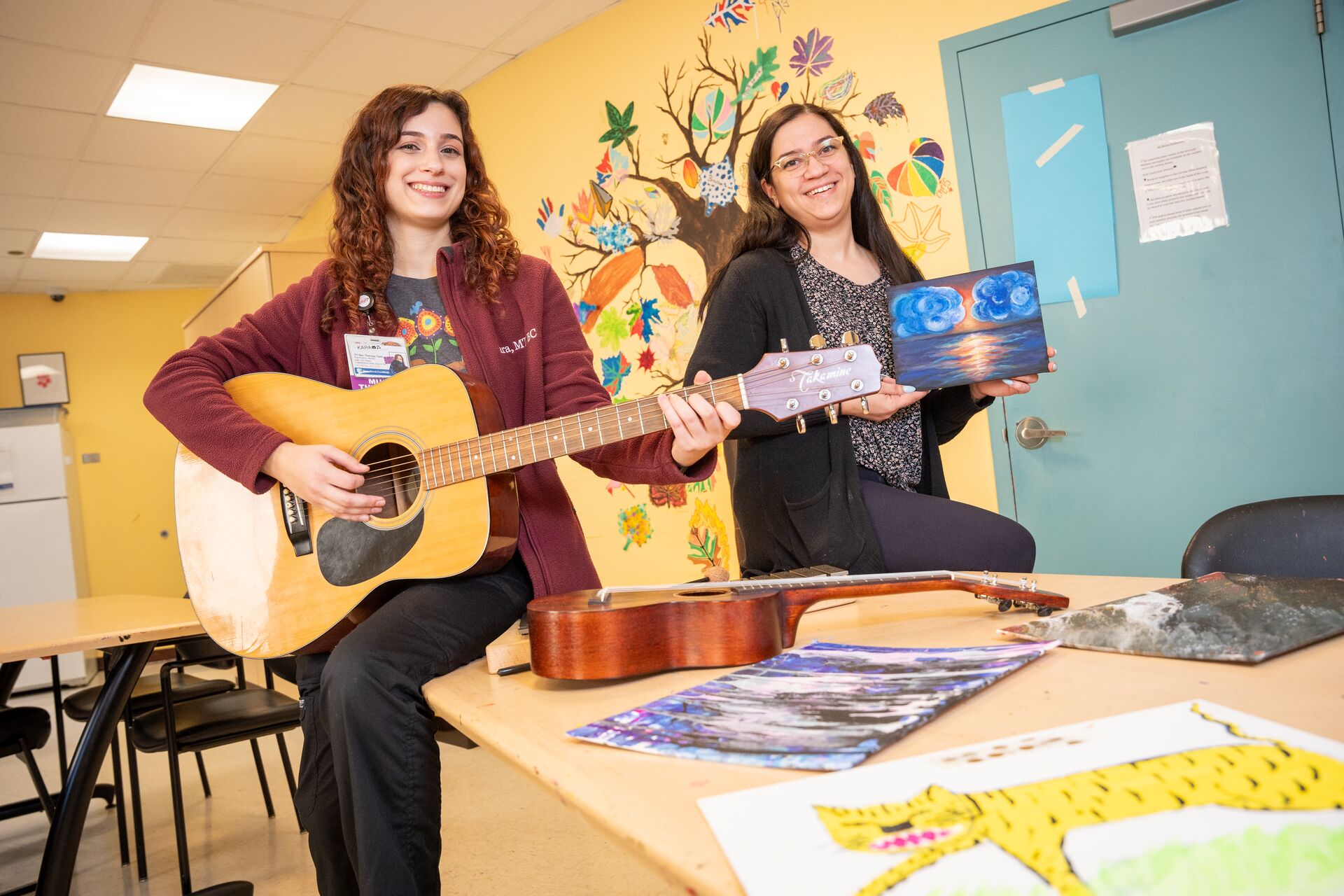 Two caucasian women in colorful art therapy room, one is playing the guitar and one is holding a small canvas