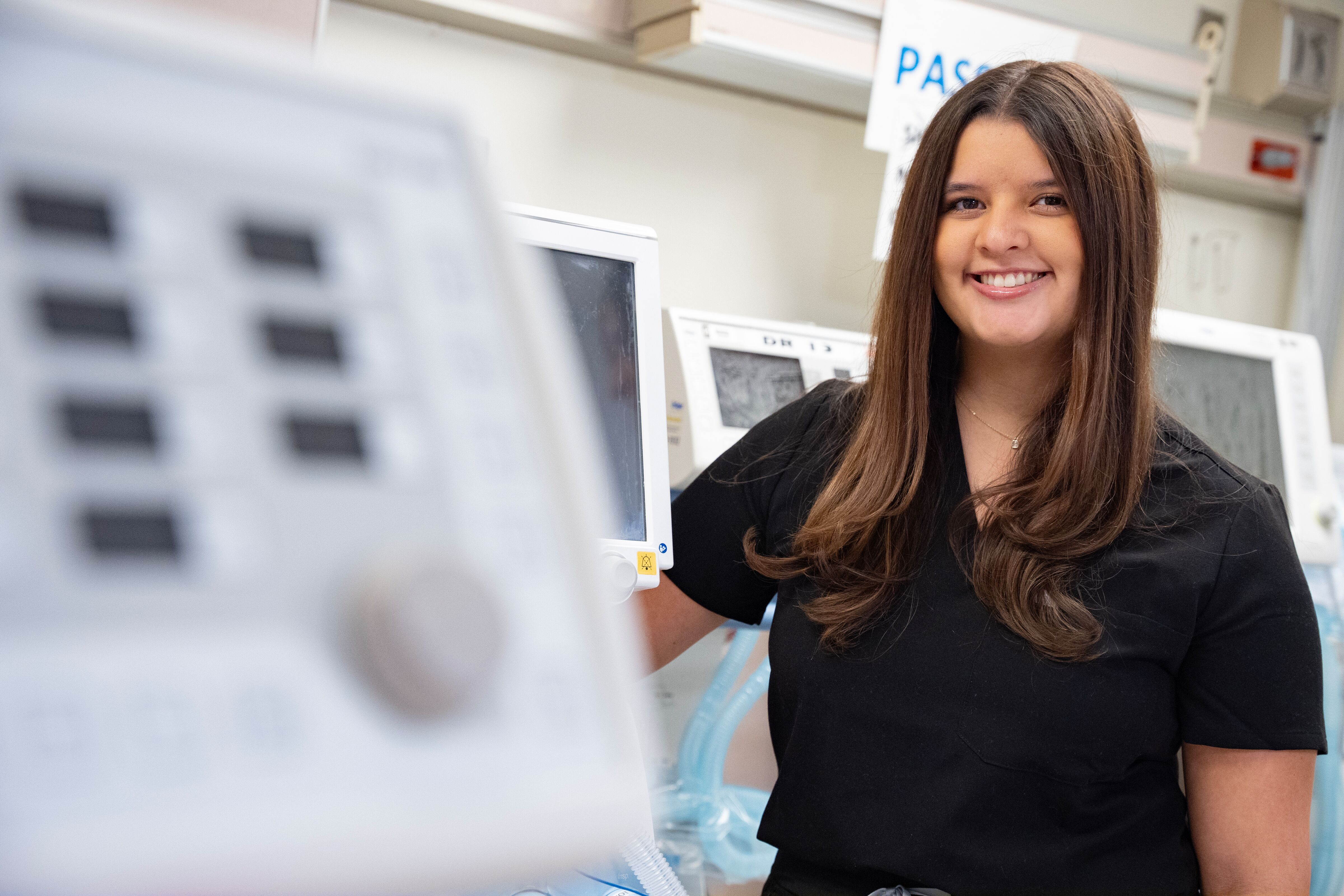 Caucasian woman with long brown hair wearing black scrubs in room with respiratory equipment