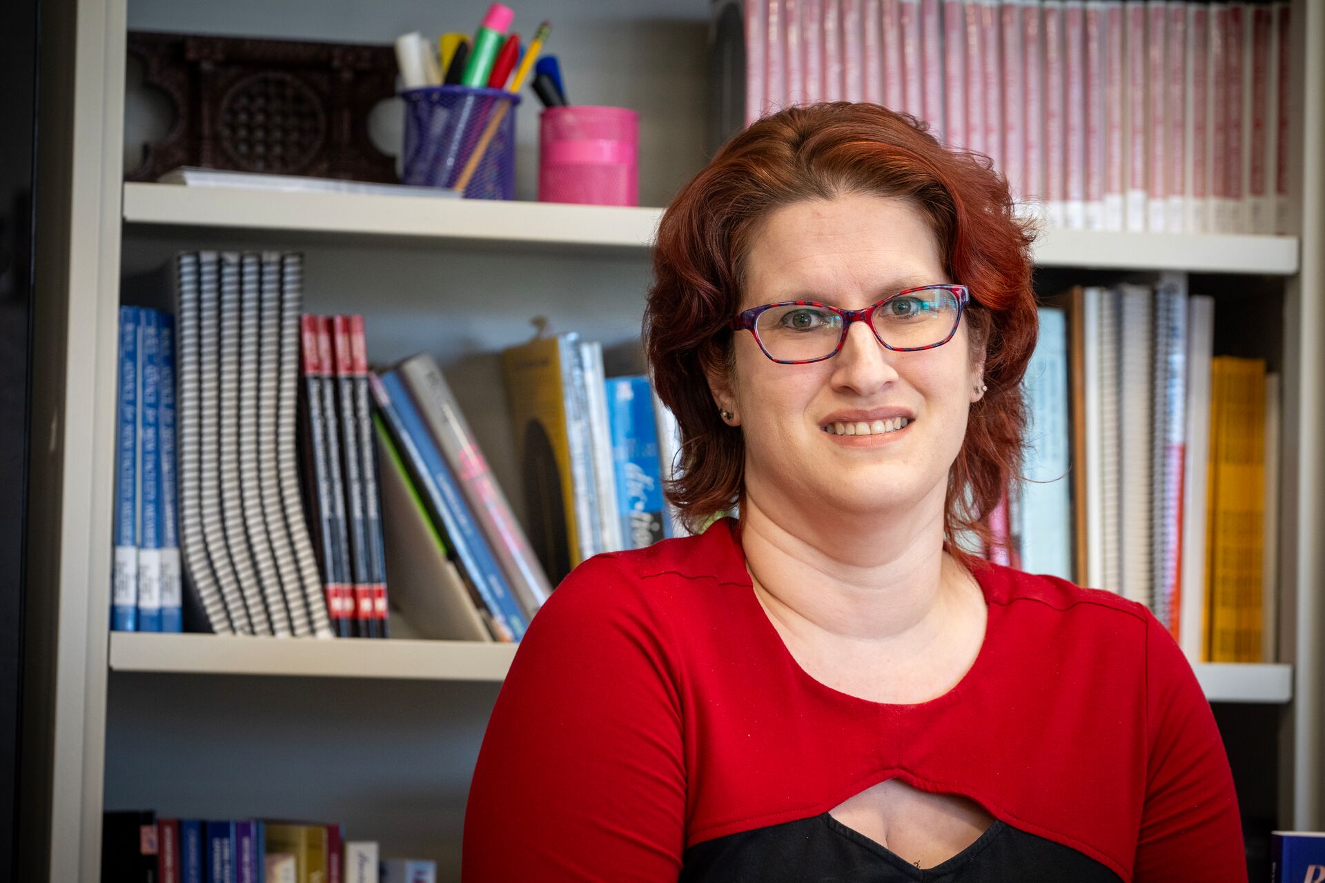 Caucasian woman with chin length bright red hair wearing black and red dress, standing in front of bookshelf