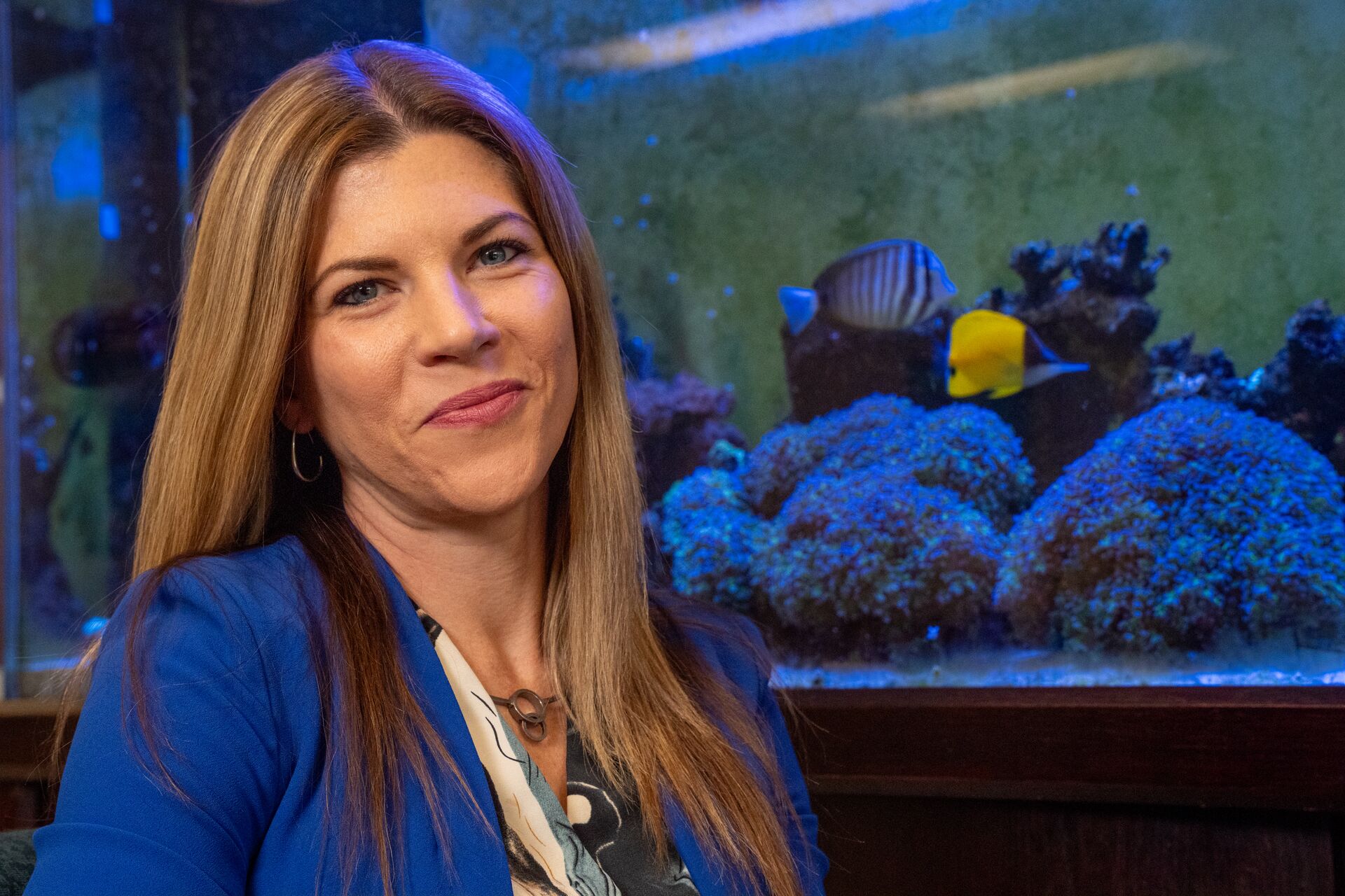 Caucasian woman with long blonde hair wearing blue blazer and patterned shirt, in front of fish tank