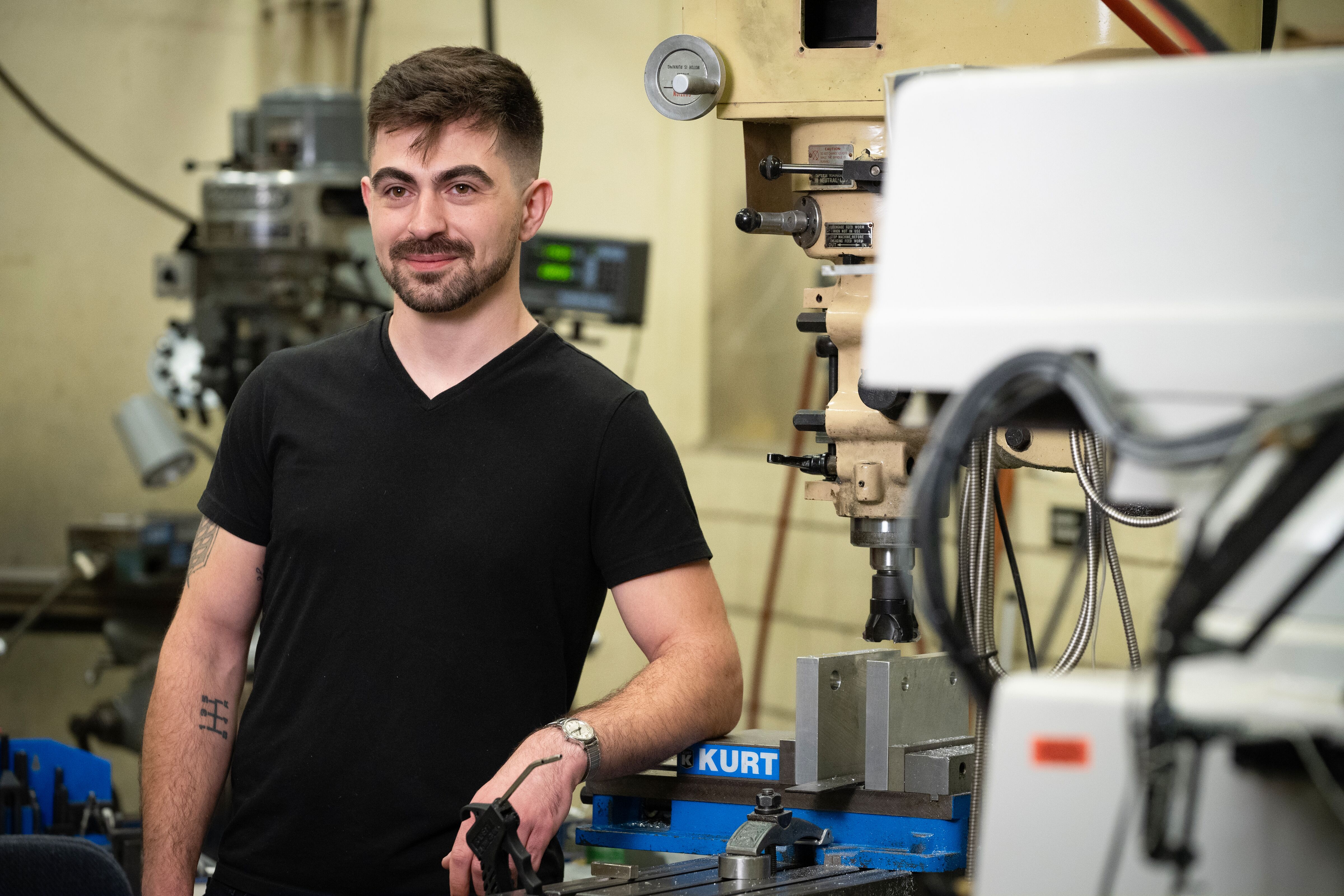 Caucasian man with short dark hair and beard and mustache, standing in a lab leaning on some machinery/equipment