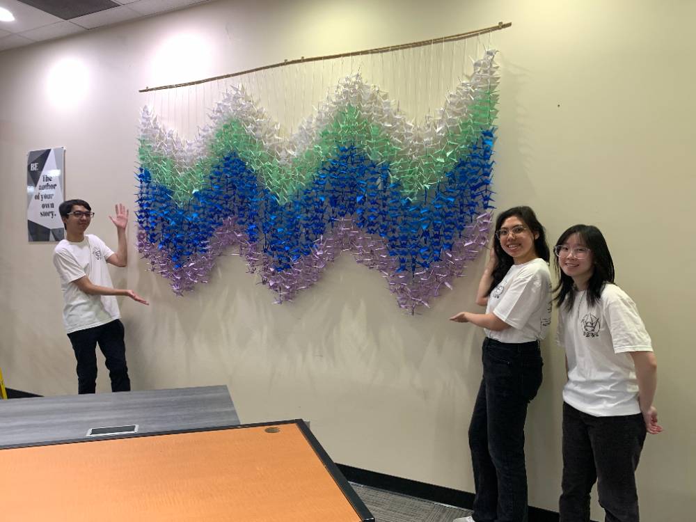 Young man and two women pointing proudly to new origami mural on the wall