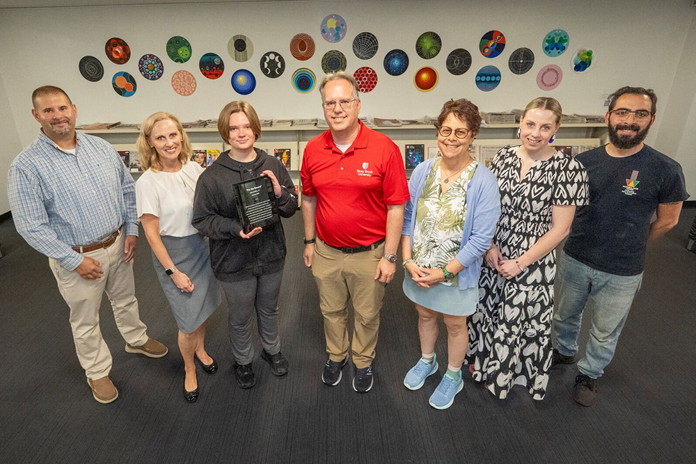 CBC members and faculty standing with student holding plaque, with the record art exhibition behind them