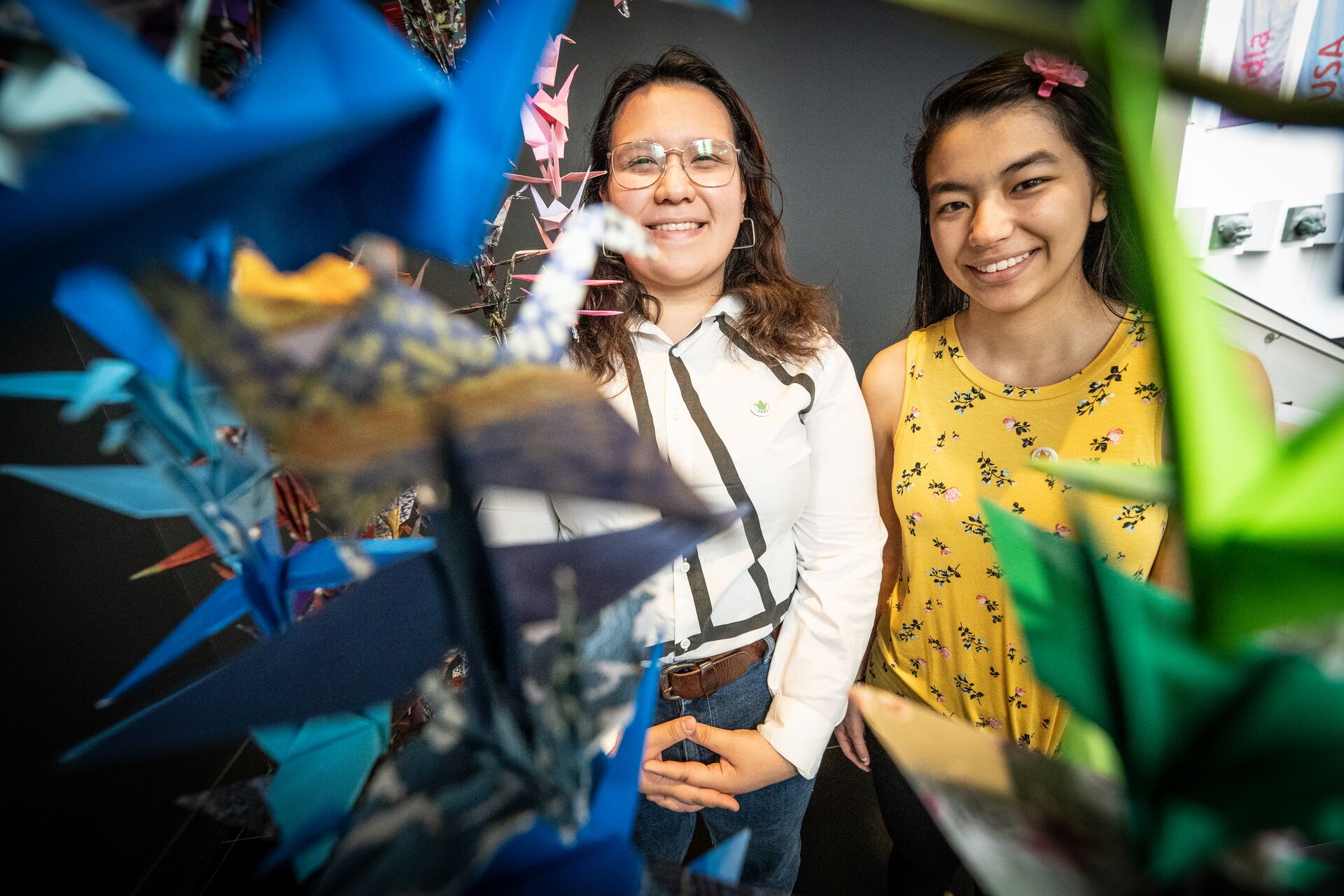 two students stood behind origami art installations that is hanging in front of the camera 