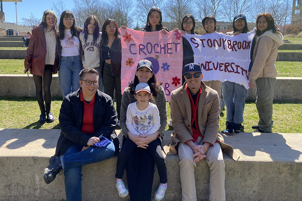 CBC members with a child sat in front of students holding up crocheted art pieces on the Staller steps 