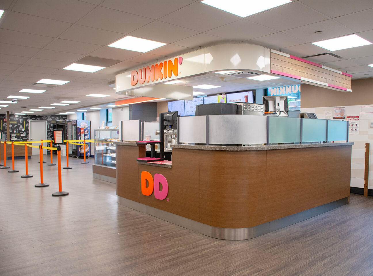 Interior view of a Dunkin' coffee shop featuring a service counter, display cases, and waiting stanchions, with a colorful logo