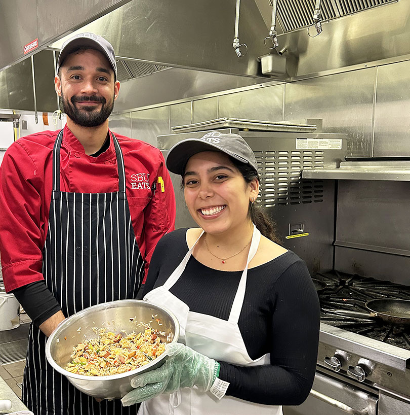 two people smiling, one holding a bowl of food, standing in a kitchen