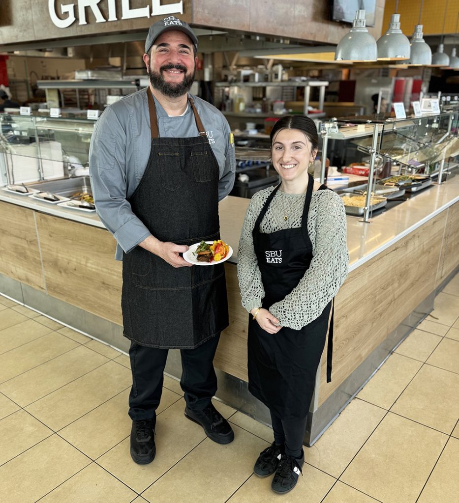 Two people in aprons, one holding a plate of food, stand in front of a serving counter