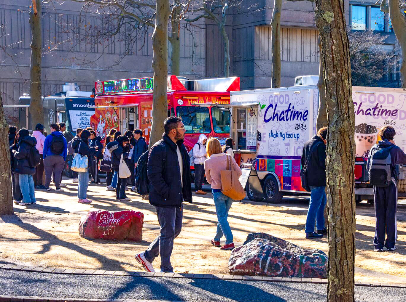 A busy outdoor scene with people gathering around food trucks, including one red truck and two others