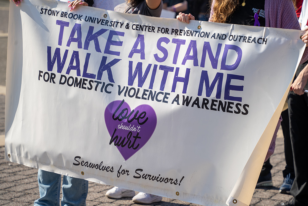 People holding a white sign with blue text that says "TAKE A STAND WALK WITH ME FOR DOMESTIC VIOLENCE AWARENESS".