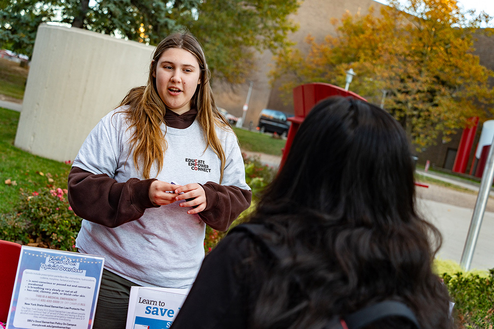 A woman in a grey sweater behind a table talking to a student with black hair outside.