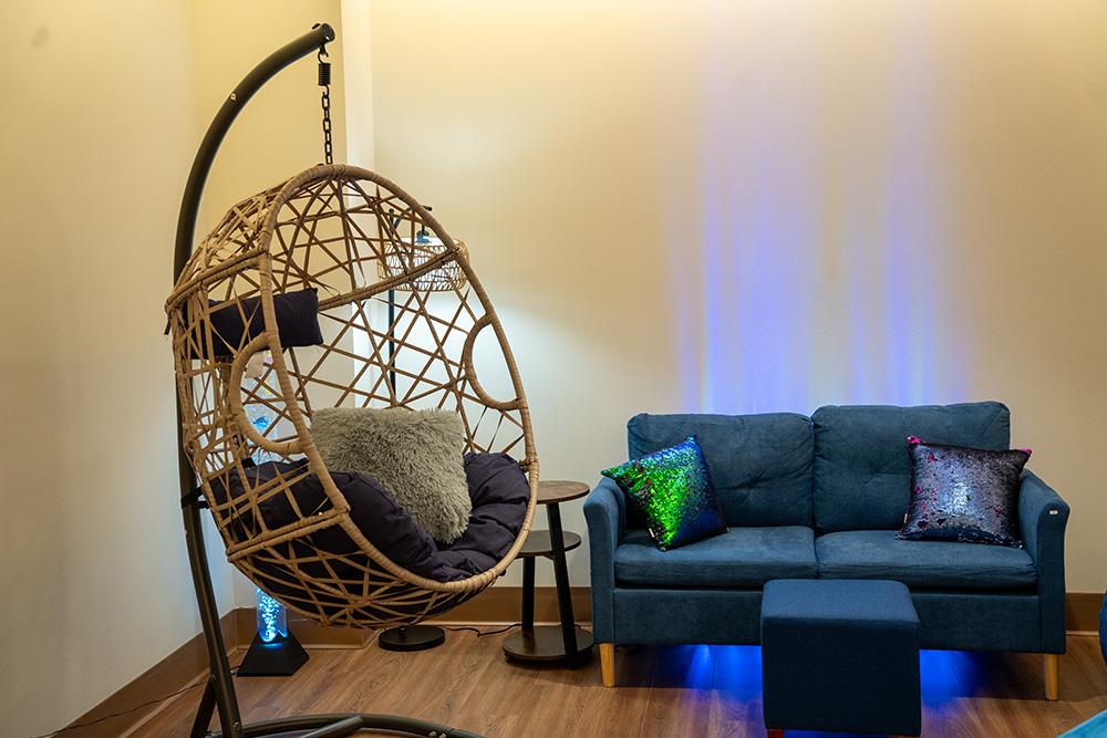 A hanging chair next to a blue couch against a white wall. Soft blue lighting is behind them.
