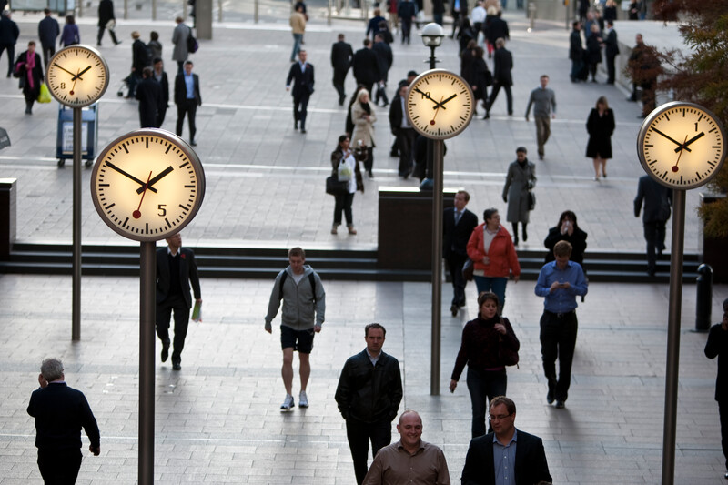 People walking past large clocks in a public plaza