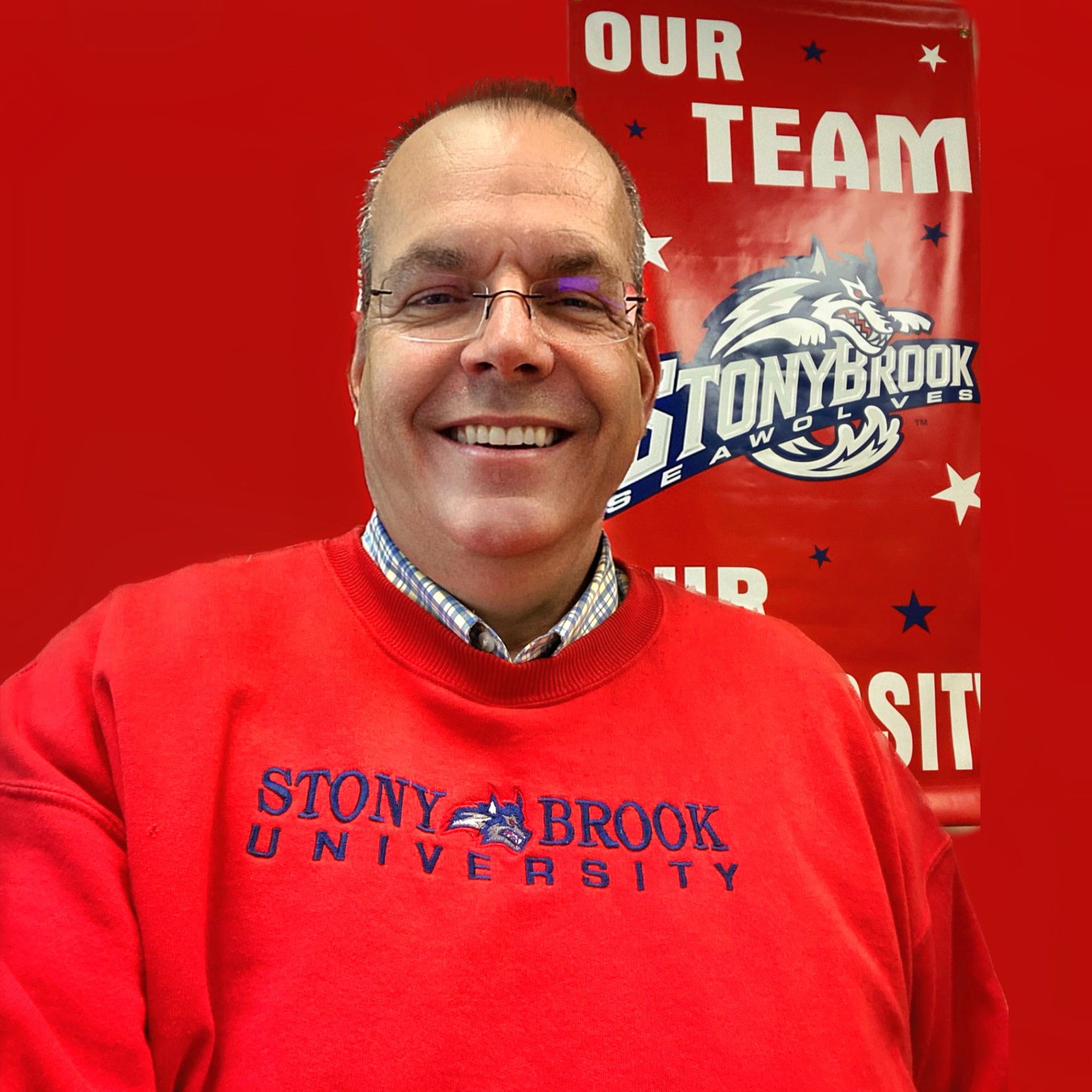 Smiling person wearing a red Stony Brook University sweatshirt, standing in front of a red Stony Brook Seawolves banner that reads ‘Our Team.