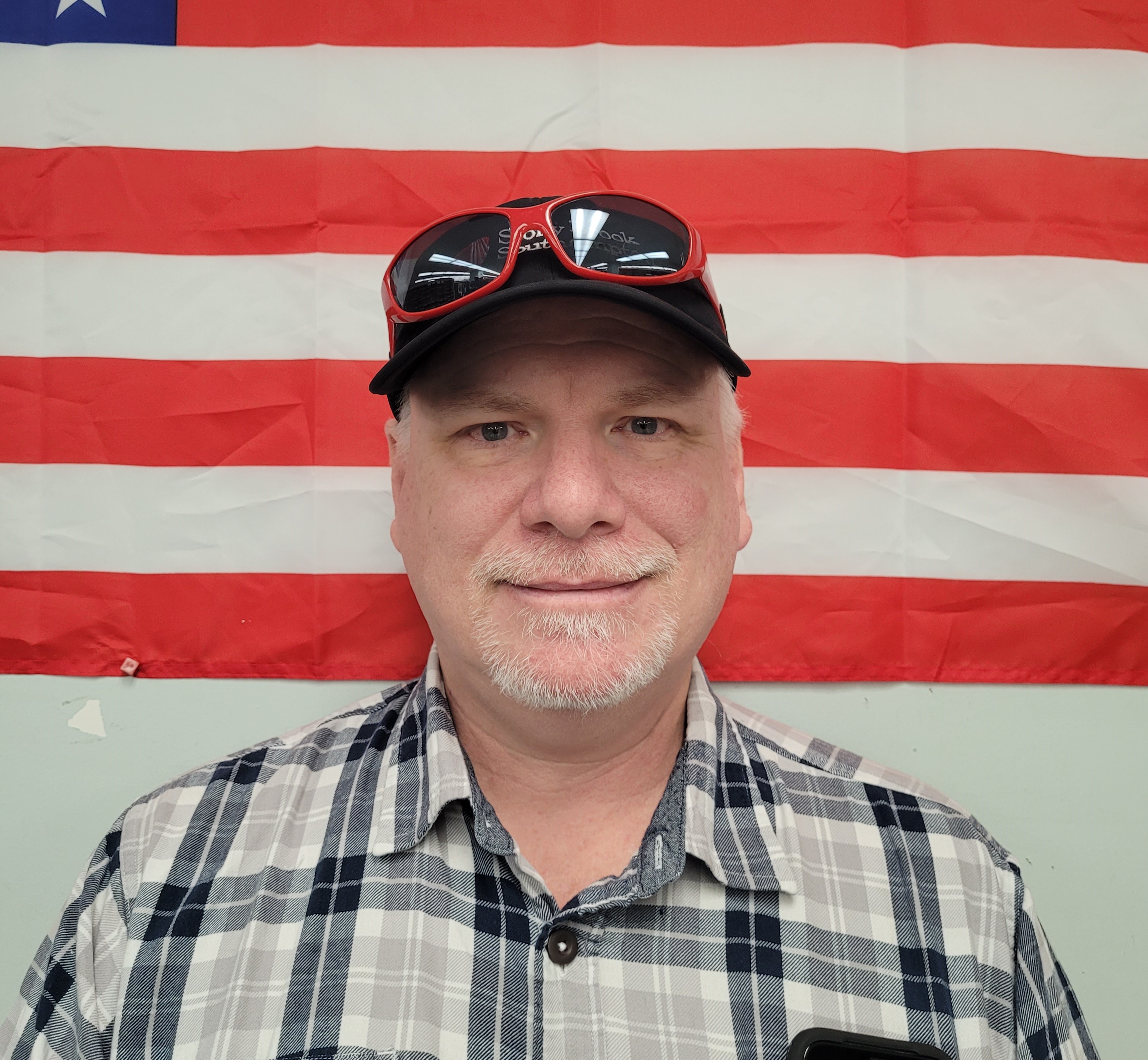 Head-and-shoulders portrait of a man wearing a black cap with sunglasses resting on the brim and a gray plaid shirt, looking at the camera with a neutral expression, standing in front of an American flag background.