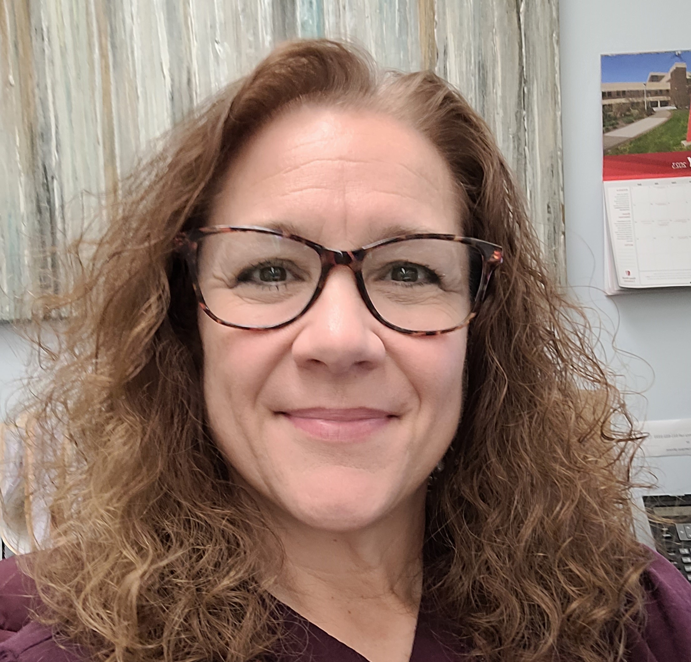 Close-up portrait of a woman with shoulder-length curly light brown hair and glasses, smiling gently, wearing a dark top, indoors with a textured wall and office items in the background.