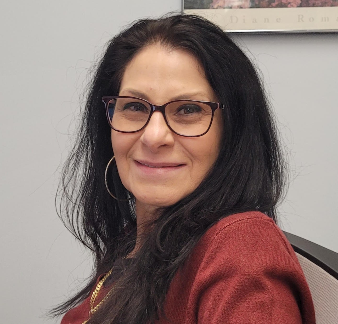 Head-and-shoulders portrait of a woman with long dark hair and glasses, smiling slightly, wearing a rust-colored top and hoop earrings, seated indoors against a light-colored wall.