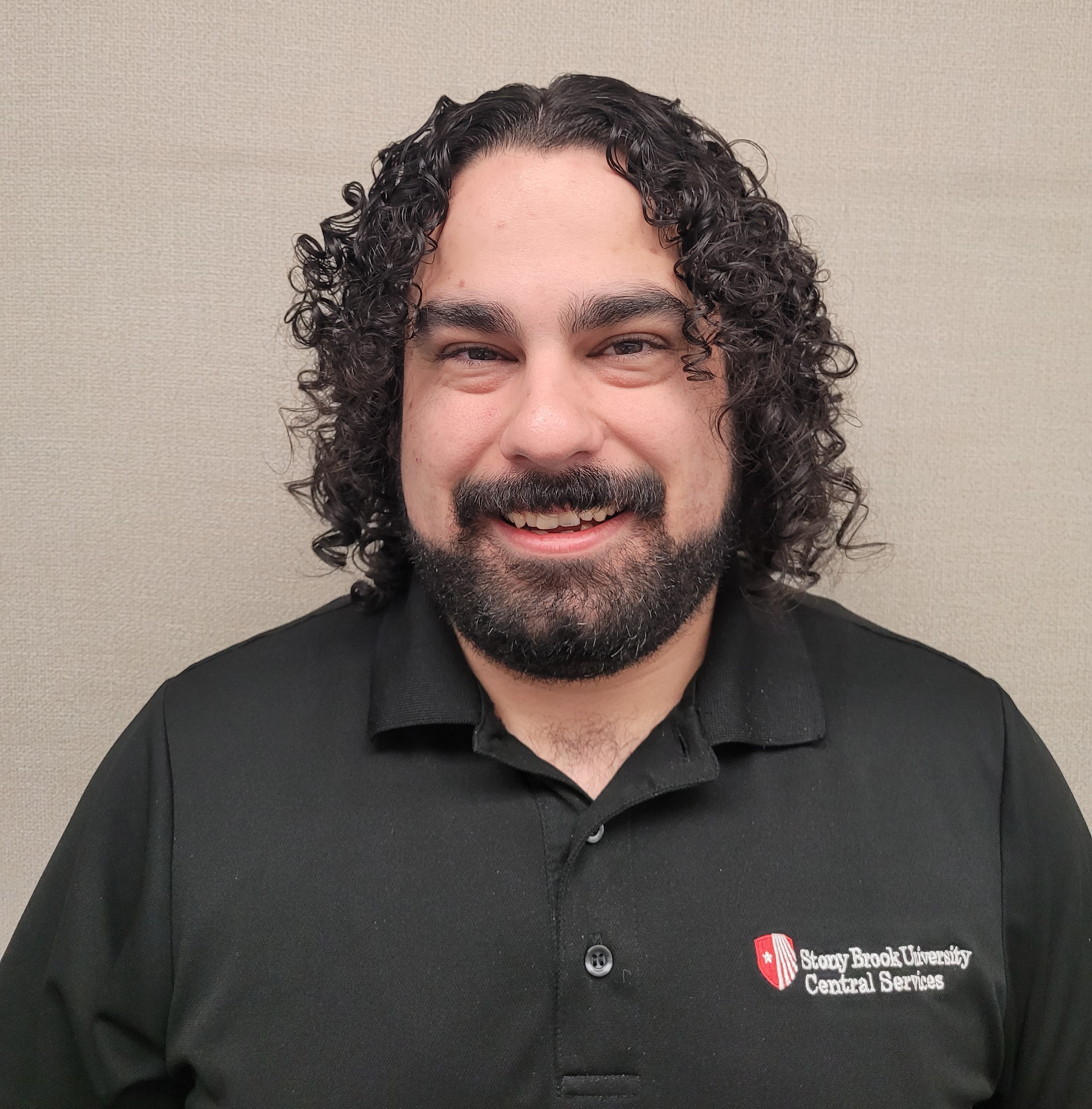 Head-and-shoulders portrait of a person with shoulder-length curly dark hair and a short beard, smiling, wearing a black polo shirt with a Stony Brook University Central Services logo, standing against a plain beige background.