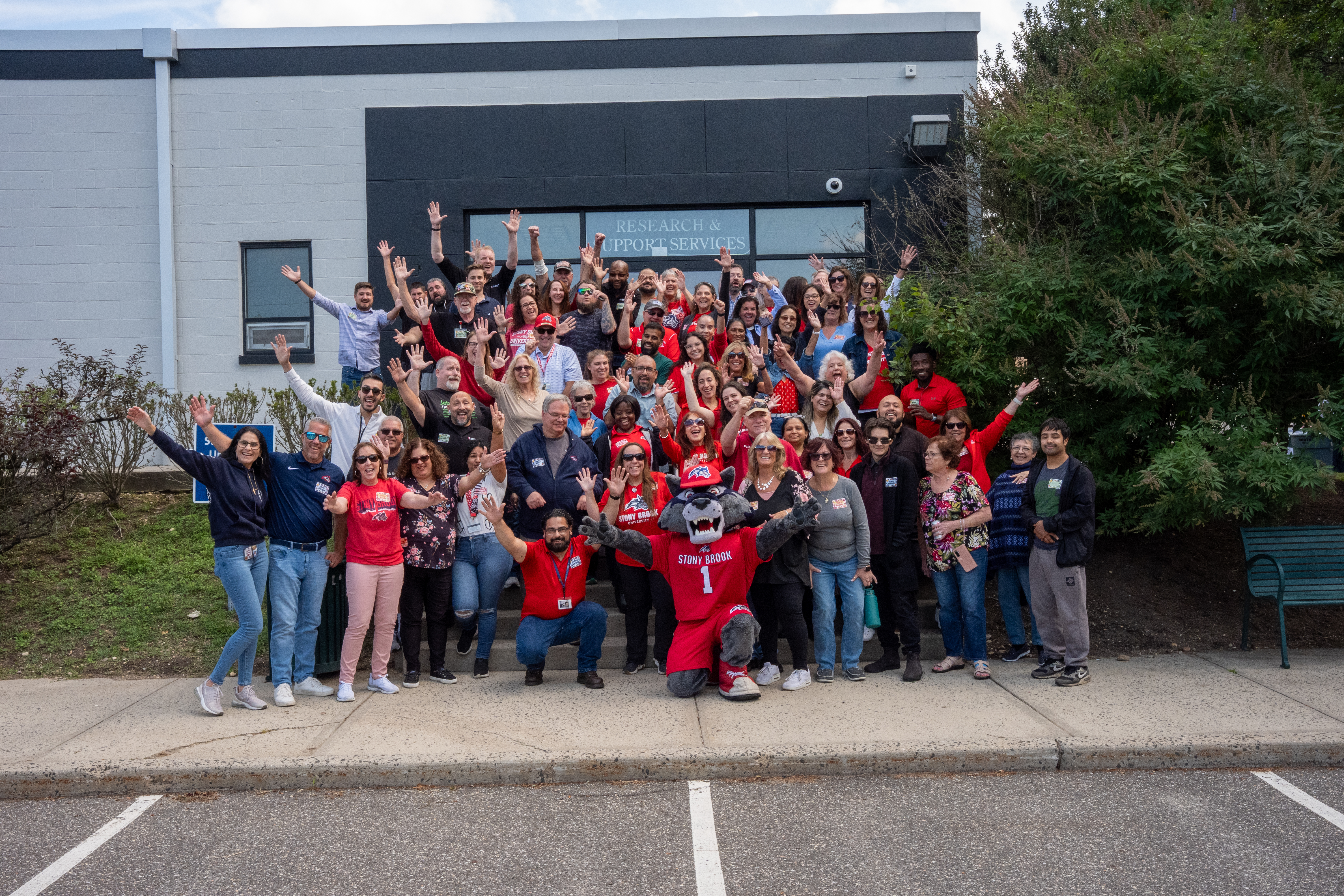 Large group posing outside a building, many wearing red and raising their arms.