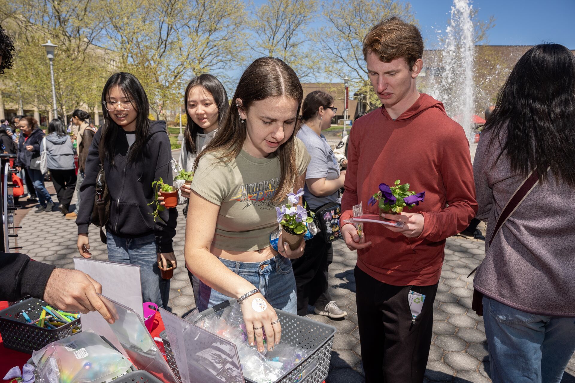 Two smiling students pose together on a paved campus plaza while holding small potted white and purple flowers.