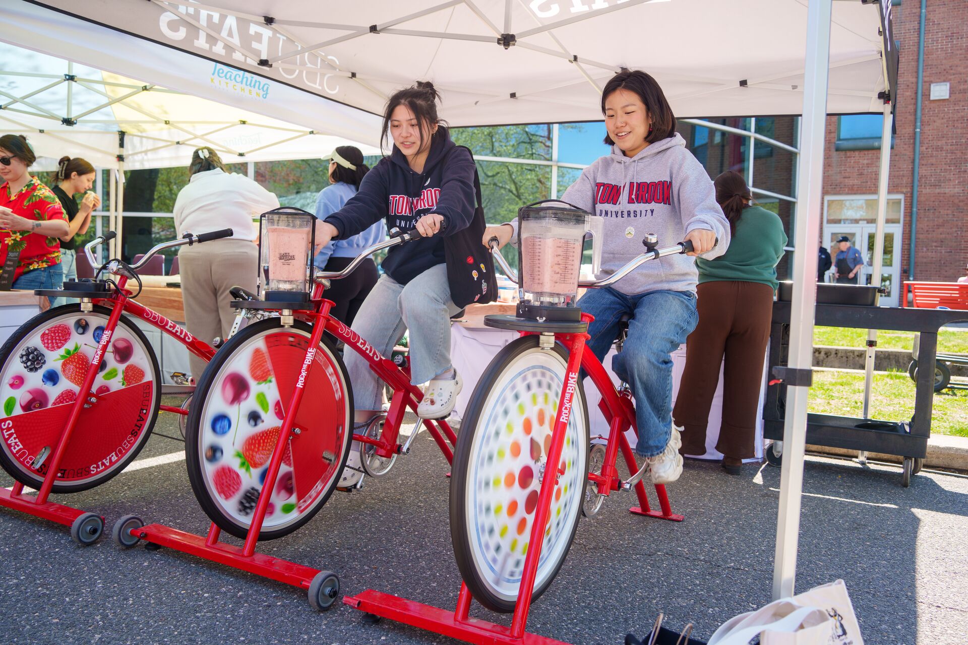 Two students ride stationary blender bikes under a campus tent, pedaling to mix smoothies in attached blender jars.