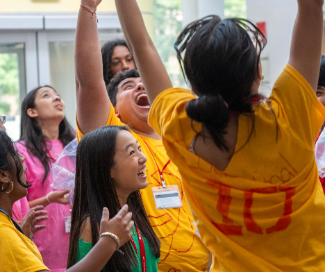 A group of diverse students are gathered indoors, looking upwards and cheering enthusiastically. Several are wearing matching yellow t-shirts, and one student in the center has their mouth wide open in a joyful shout with arms raised high.