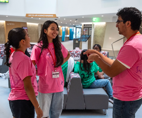 In a well-lit campus lobby, three students wearing bright pink t-shirts and name tags stand in a circle talking. One student on the right is gesturing with his hands while speaking to two female students who are listening intently. Other students sit on modern grey sectional couches in the background.