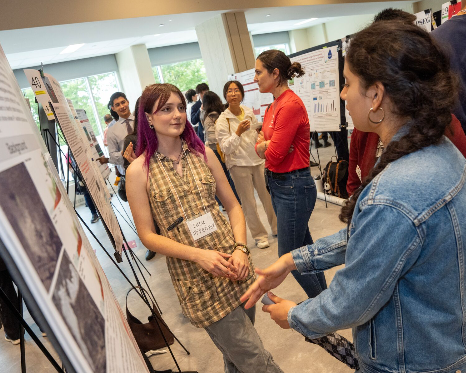 An indoor academic poster session where a student wearing a plaid dress and a name tag stands next to her display board. She is engaged in conversation with another student in a denim jacket who is gesturing toward the research poster. Other attendees and display boards fill the room.