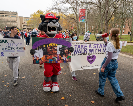 A campus awareness walk moving down an asphalt path lined with trees. In the center, a person in a grey and red wolf mascot costume holds a colorful "Seawolves for Survivors" sign. Other students walk alongside, holding a megaphone and a large white banner advocating for domestic violence awareness.
