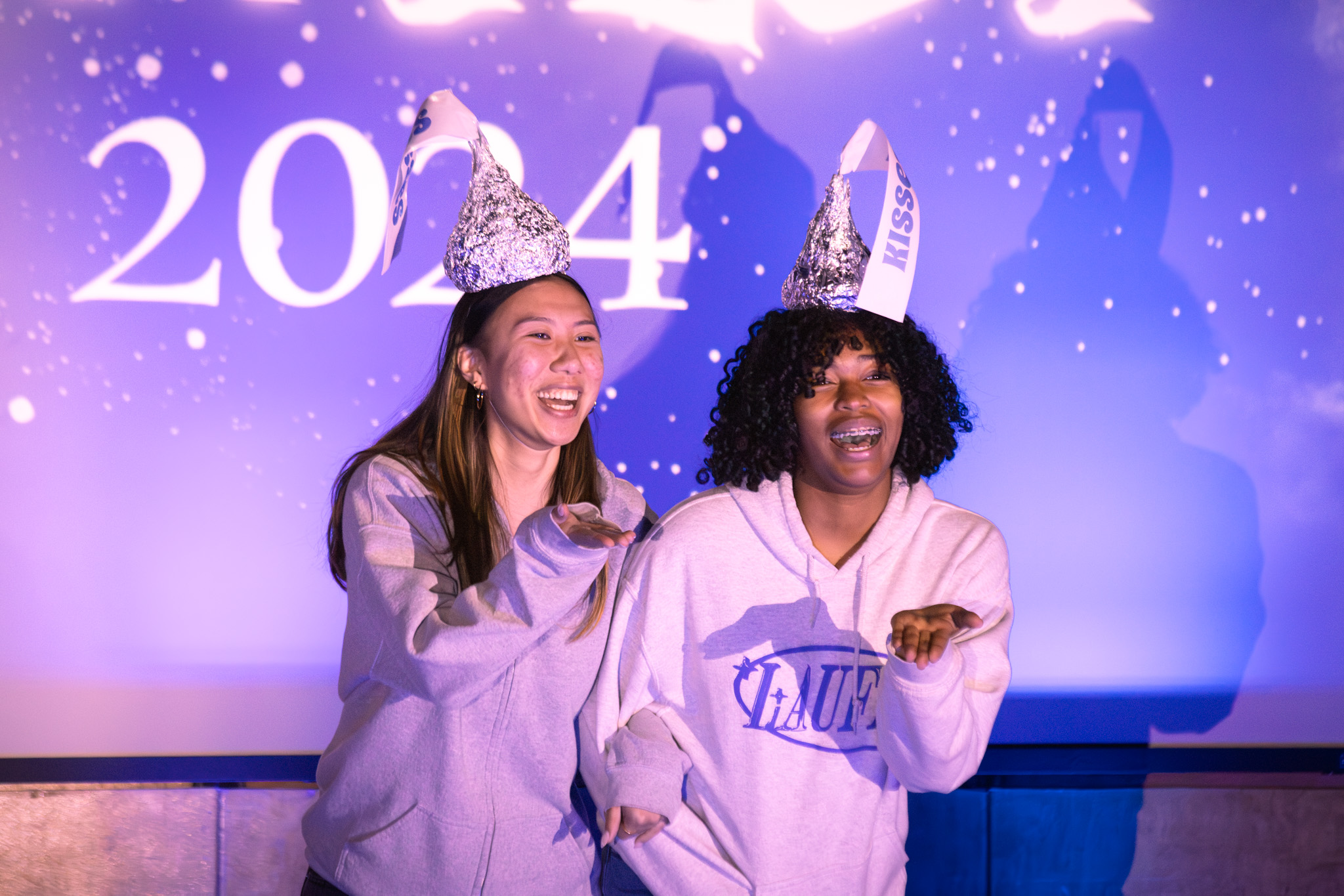 Two individuals wearing silver party hats are joyfully posing in front of a purple backdrop with the year "2024" displayed.