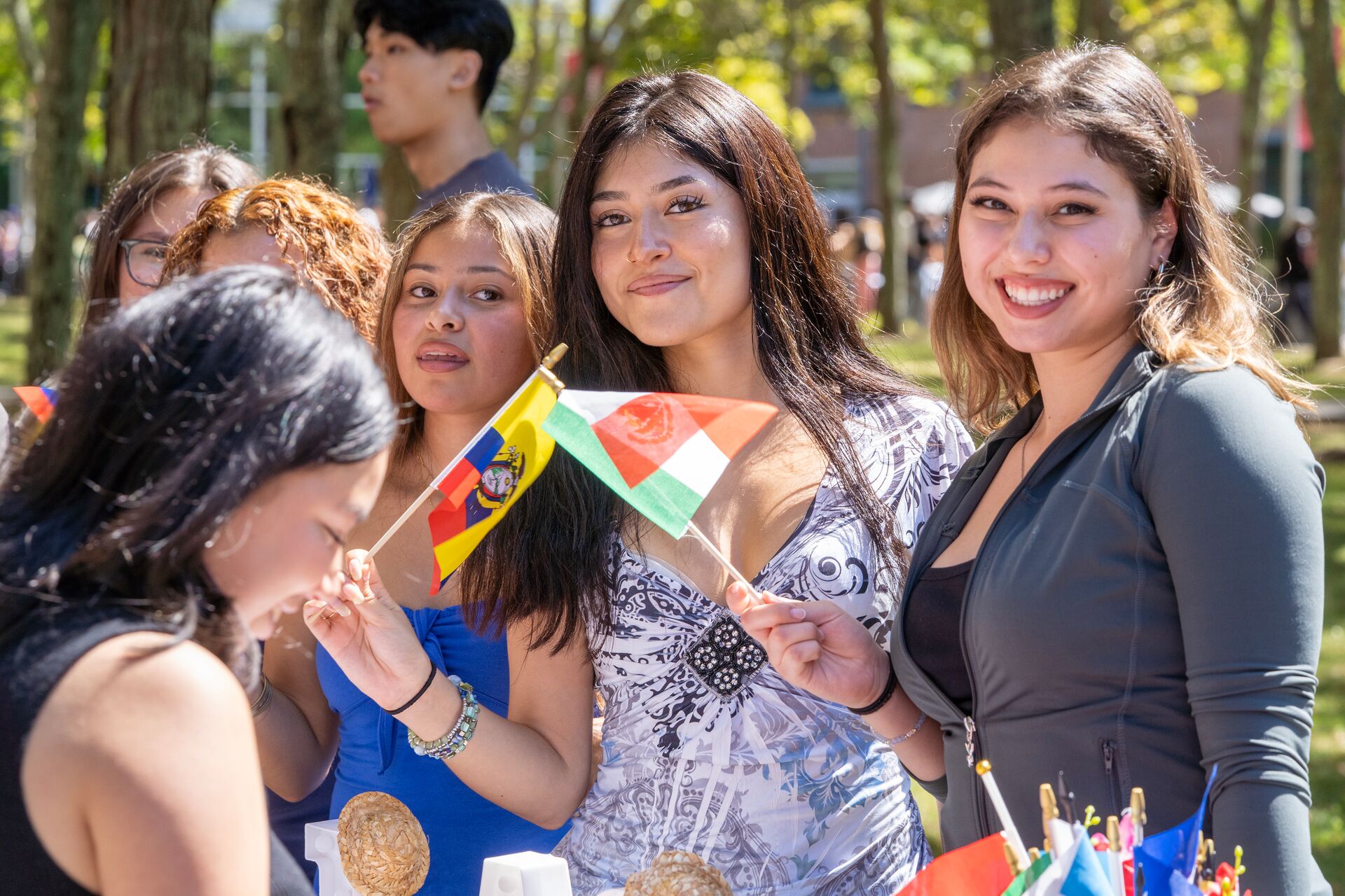 Four young women stand outdoors, smiling and holding small national flags at an event, with trees and other people visible in the background.