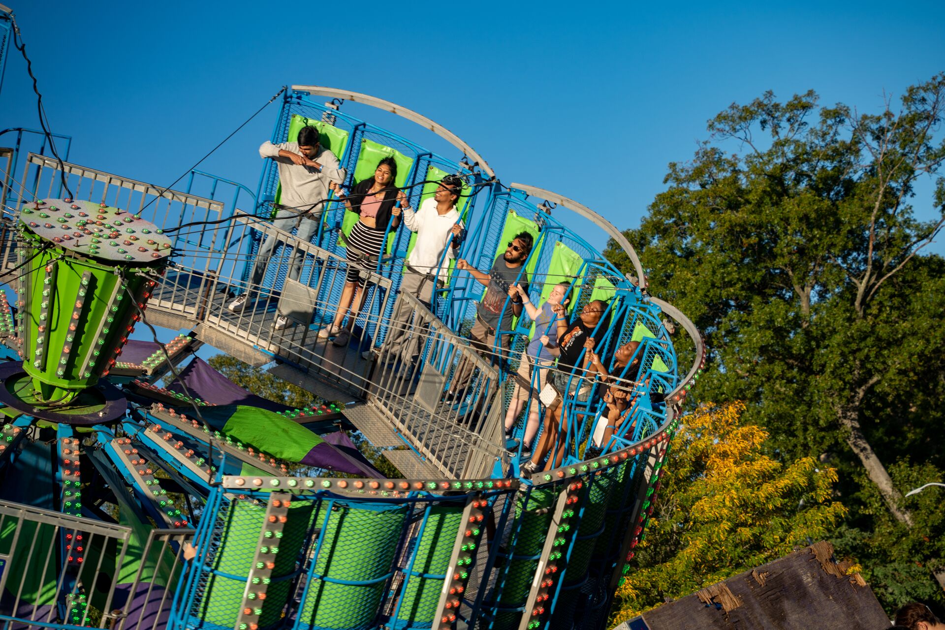 Group of five people enjoying a carnival ride with colorful lights and structures against a clear blue sky.