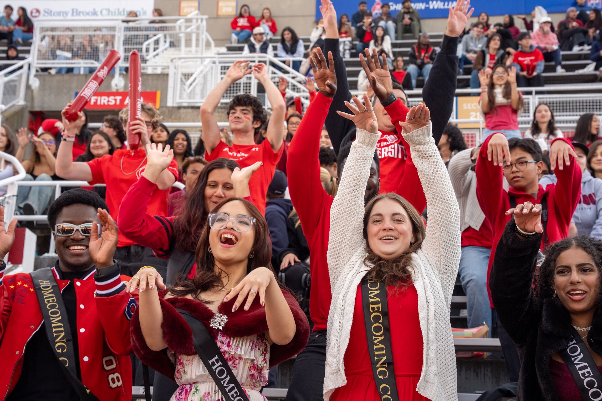 A diverse group of students in red attire celebrating at a homecoming event, waving hands and cheering in a stadium.