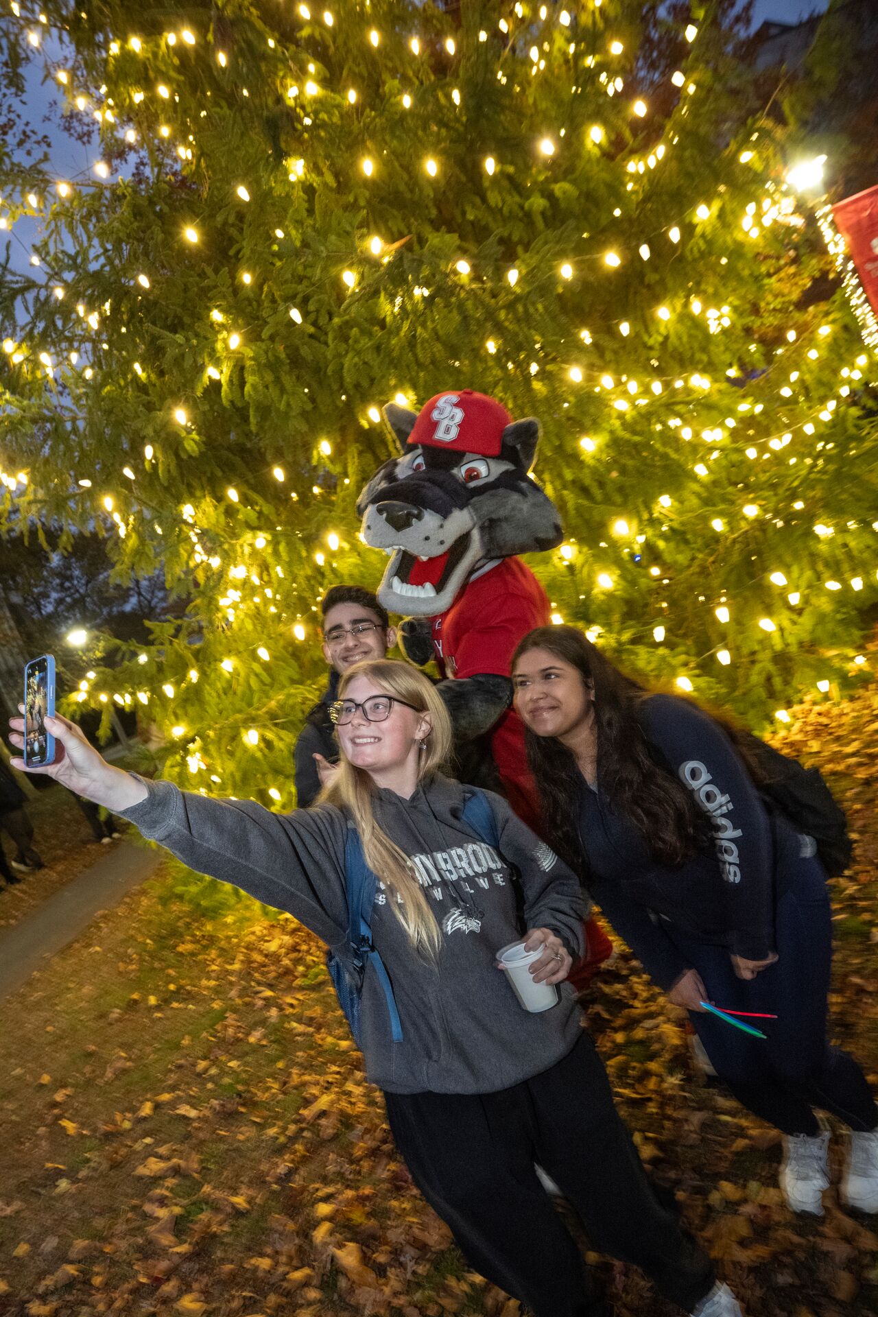Three students take a selfie with a wolf mascot in front of a large outdoor Christmas tree decorated with yellow lights.