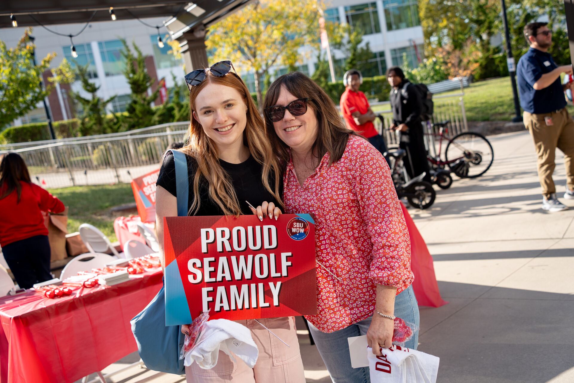 Two women pose together holding a sign that reads 'PROUD SEAWOLF FAMILY' at an outdoor event with tables and attendees in the background.