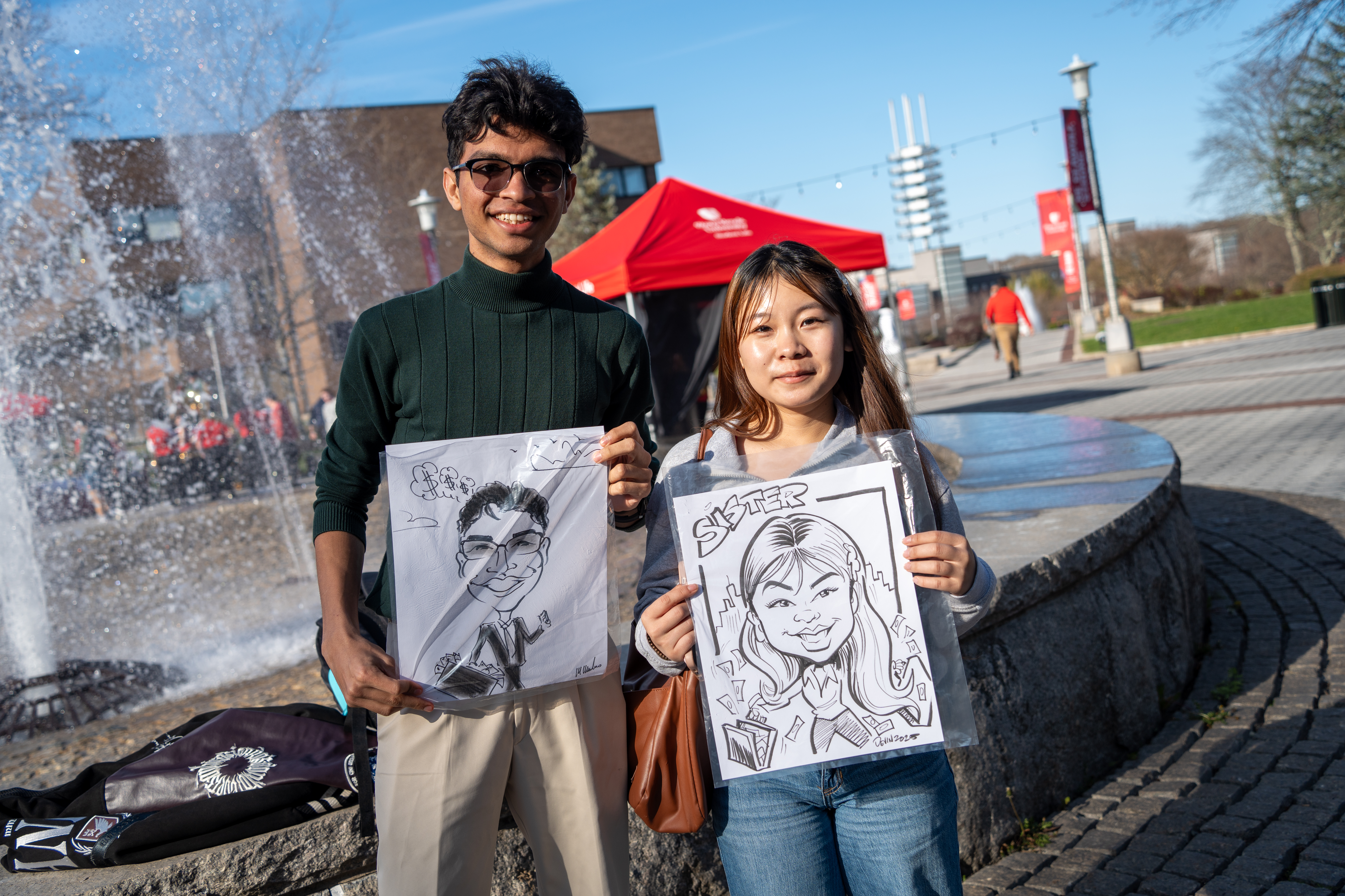 Two smiling university students stand outdoors near a fountain, each proudly holding up a personalized black-and-white caricature drawing of themselves in protective plastic sleeves.